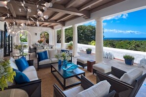 A covered outdoor seating area with white and blue furniture, a glass coffee table, and a view of the ocean. - Villa Elsewhere (Holetown)