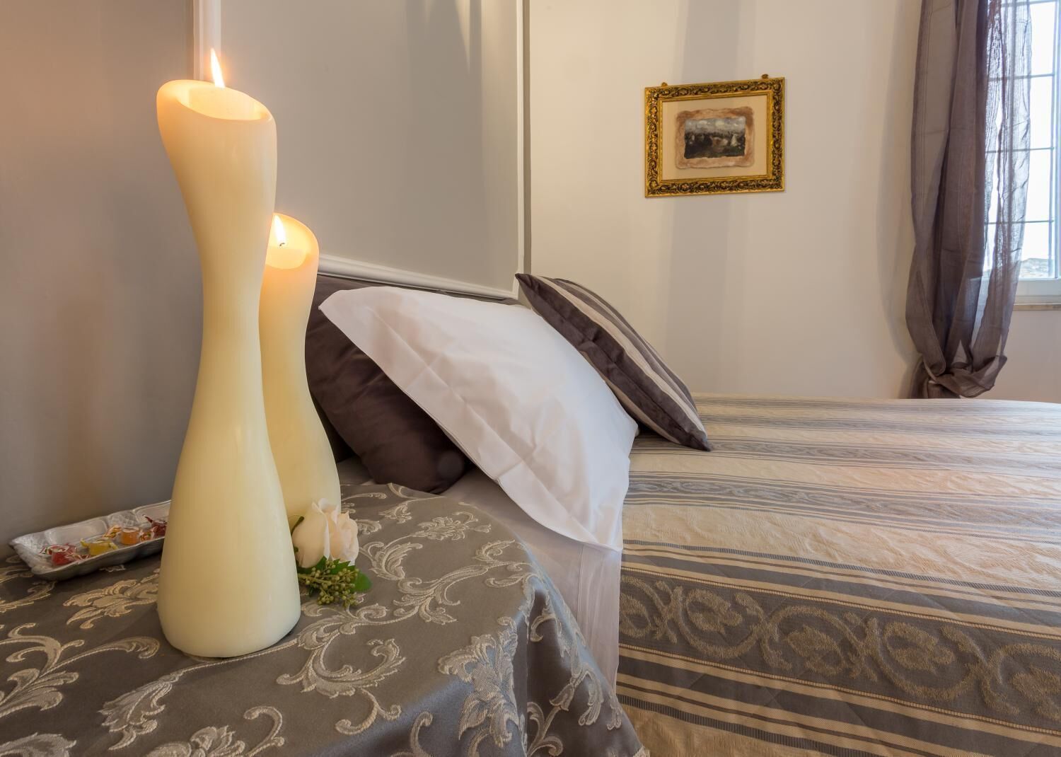 Una camera d'albergo con un letto, due grandi candele, un quadro appeso al muro e una finestra con tende.