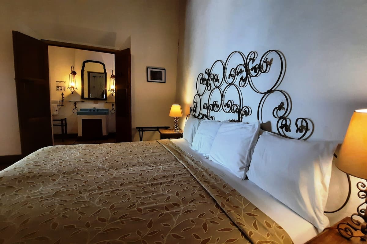 Ein Hotelzimmer mit Bett, Nachttisch mit Lampe, sichtbarem Badezimmer hinter einer offenen Tür und einem dekorativen Wandvorhang als Kopfteil.