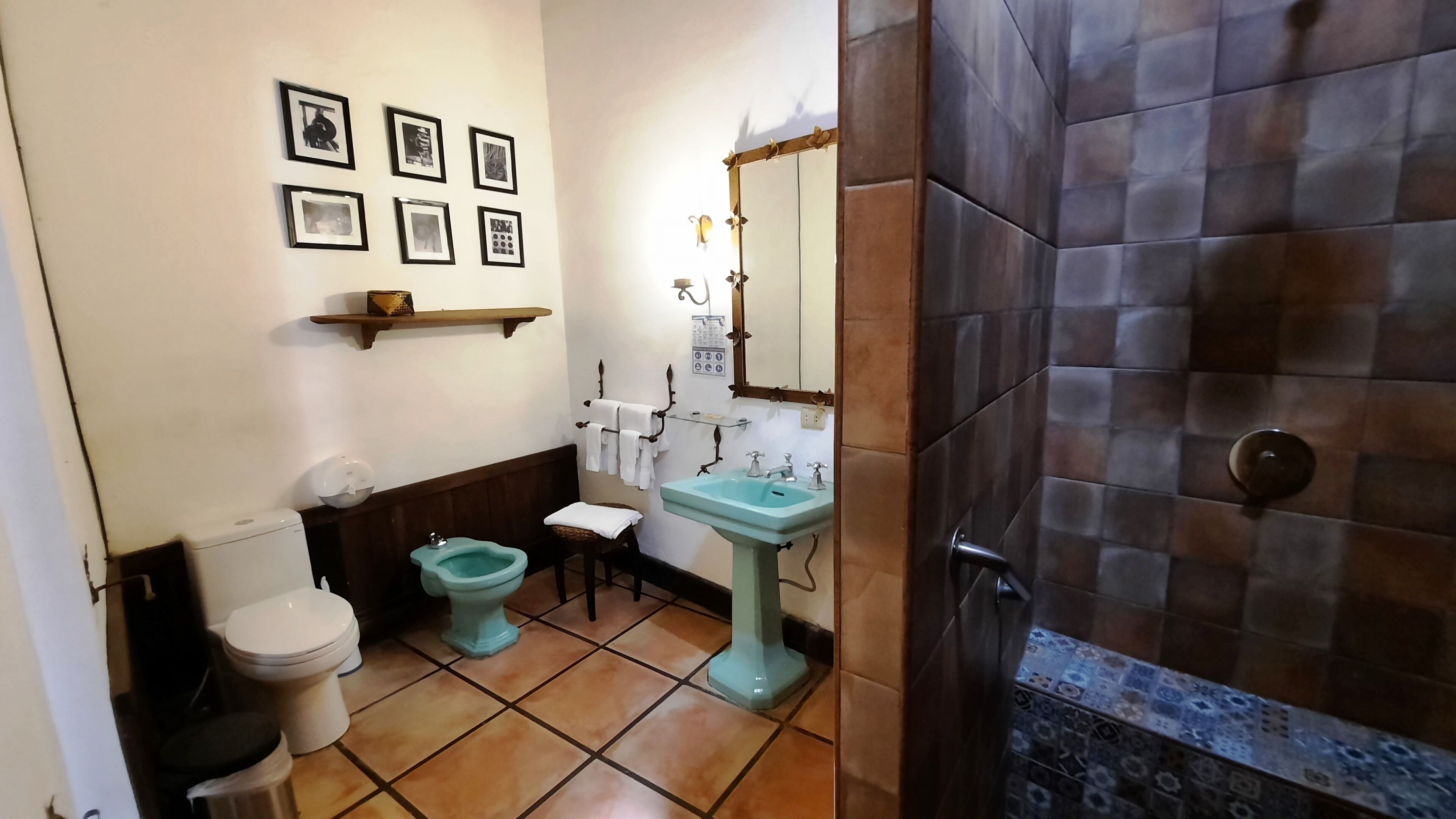 Un baño con inodoro, bidé, lavamanos y espejo. Hay cuadros enmarcados en la pared y un estante sobre el lavamanos.