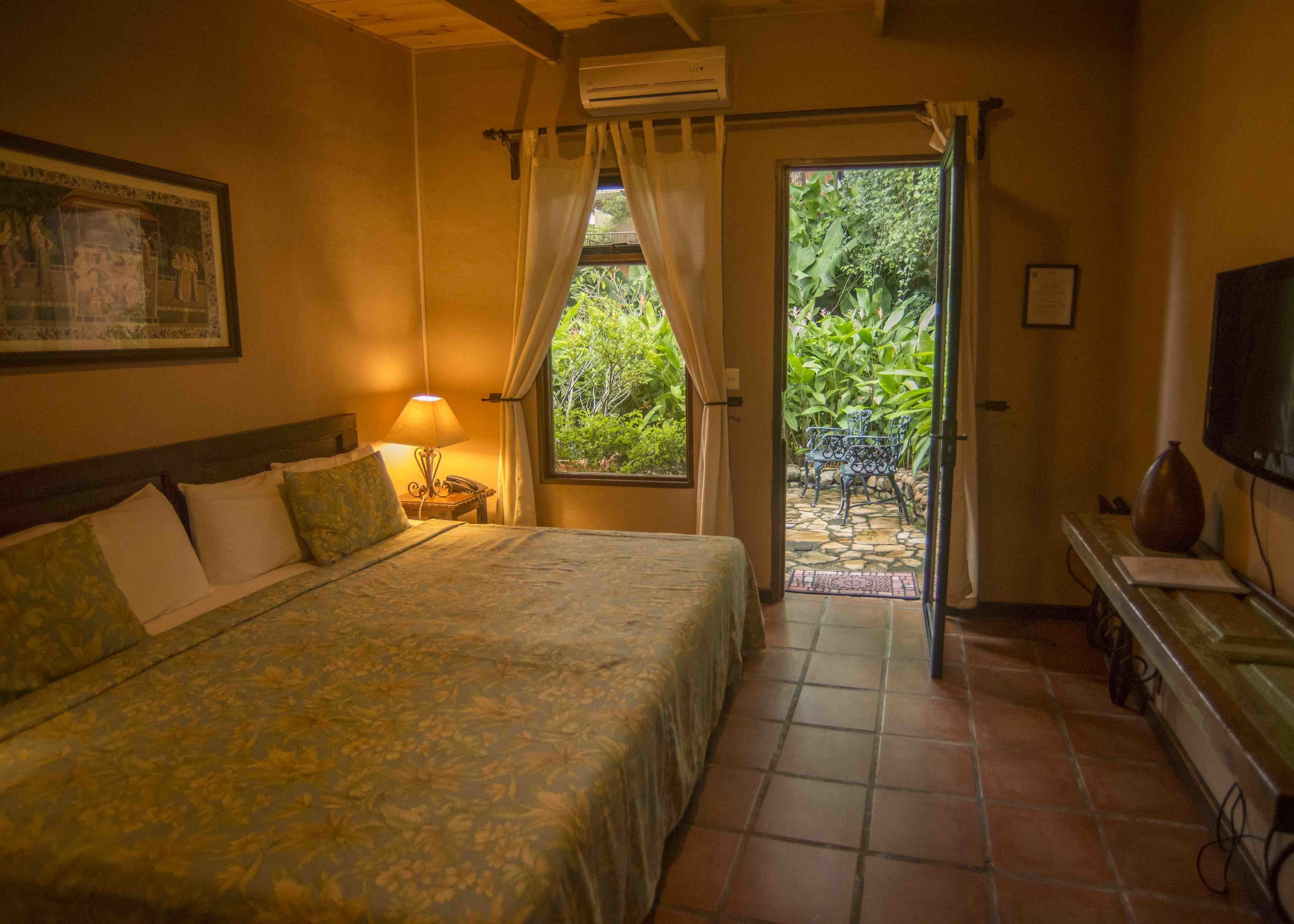 Un dormitorio con cama, mesita de noche, lámpara, aire acondicionado, televisor y vista a un jardín a través de una puerta.