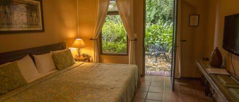 Un dormitorio con cama, mesita de noche, lámpara, aire acondicionado, televisor y vista a un jardín a través de una puerta.