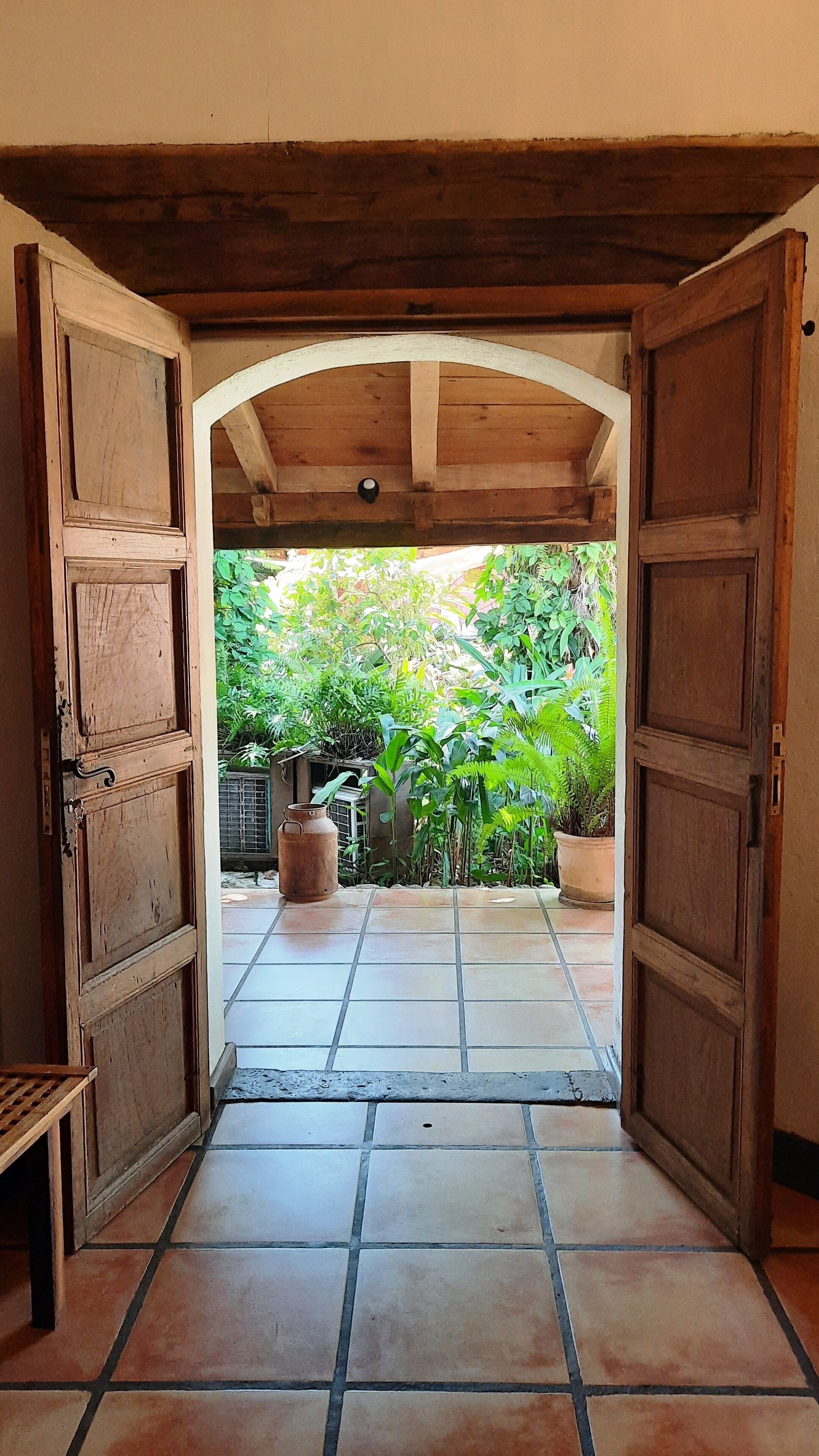 Uma porta de madeira aberta dá para um pátio com azulejos, plantas em vasos e vista de um verde exuberante.