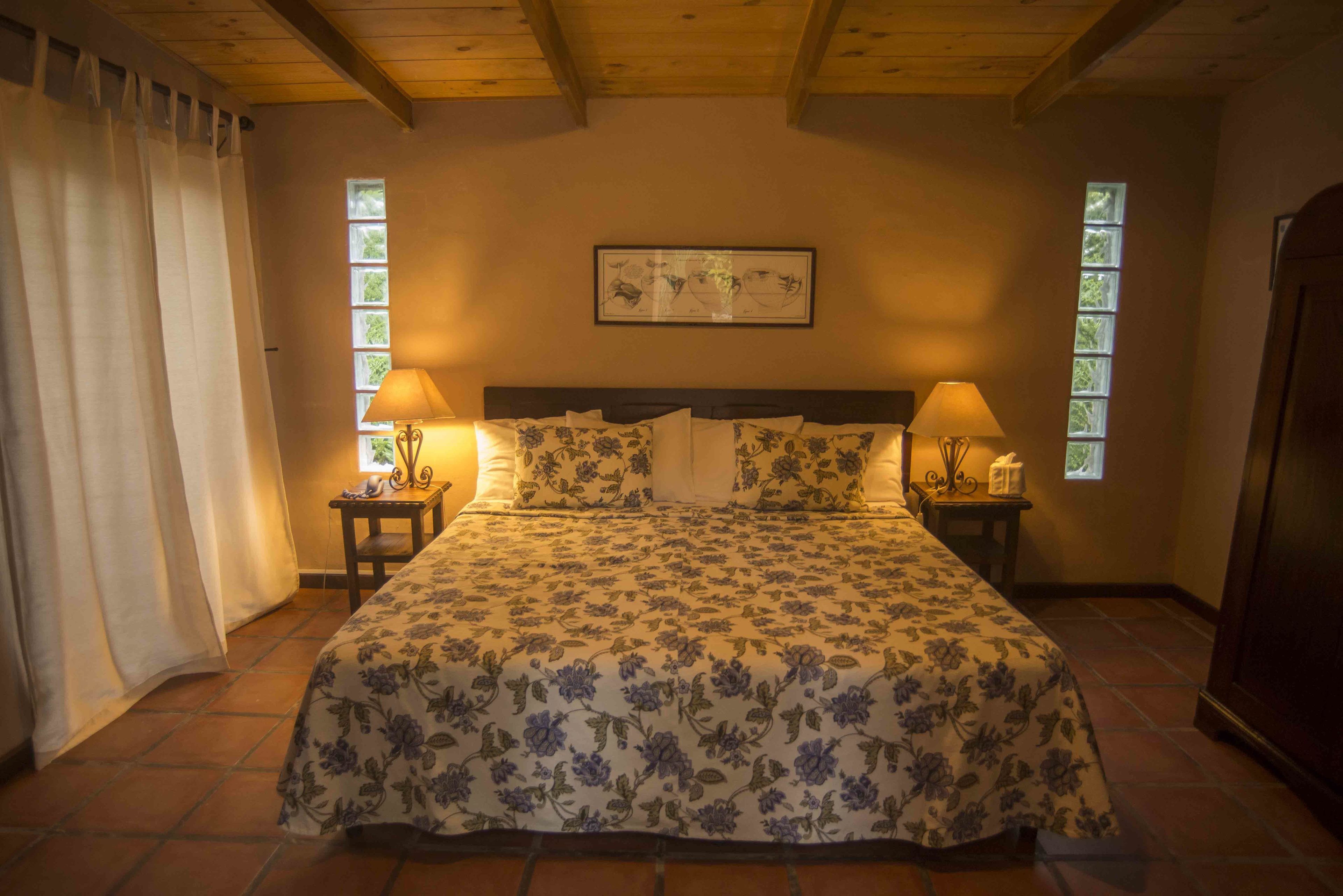A bedroom with a floral bedspread, two bedside lamps, a framed artwork on the wall, and a window on the right side.
