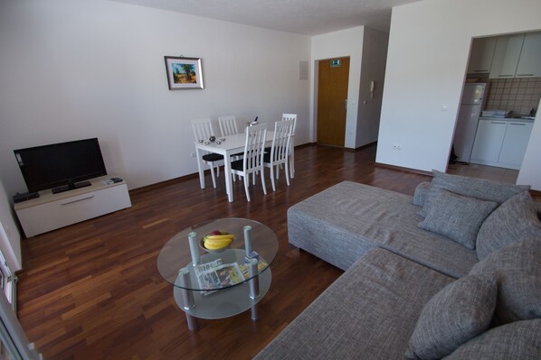 A modern living room with a grey sofa, a glass coffee table with magazines, a dining area with white chairs, a TV on a low cabinet, and a kitchen area with white cabinets. - Apartments Kelic (Okrug)
