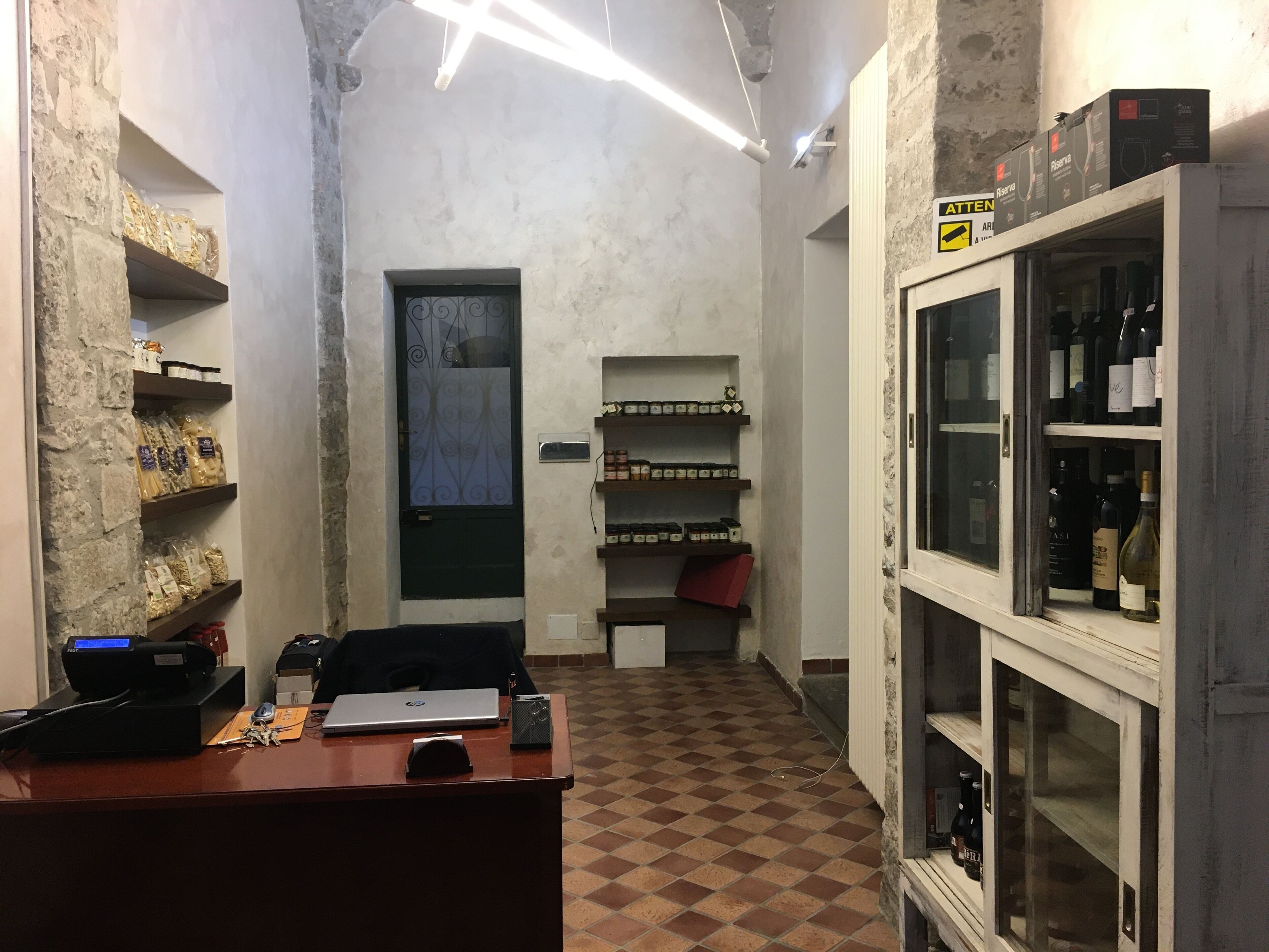A narrow hallway with a checkered floor, a desk with a laptop, a printer, and a security camera. Shelves on the left hold packaged goods, and a wine rack on the right displays bottles.