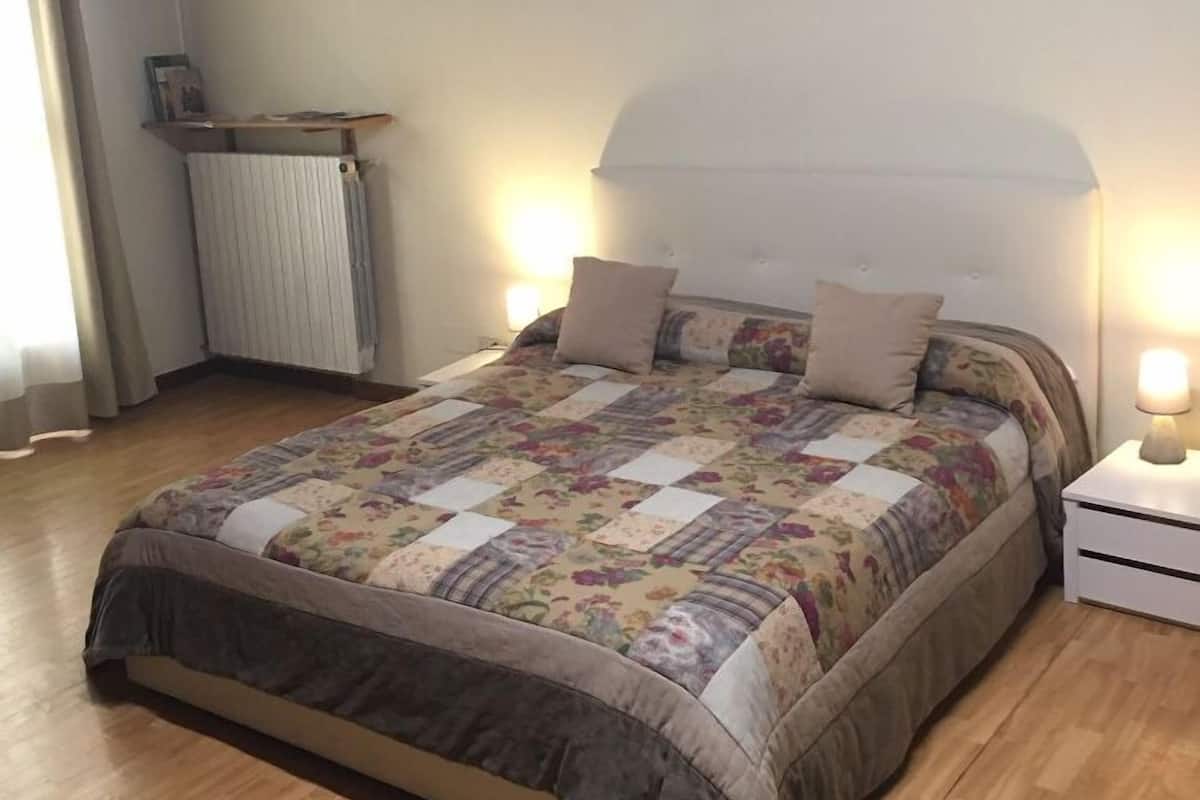 A bedroom with a wooden floor, a bed with a floral quilt, a bedside lamp, and a wall-mounted picture.