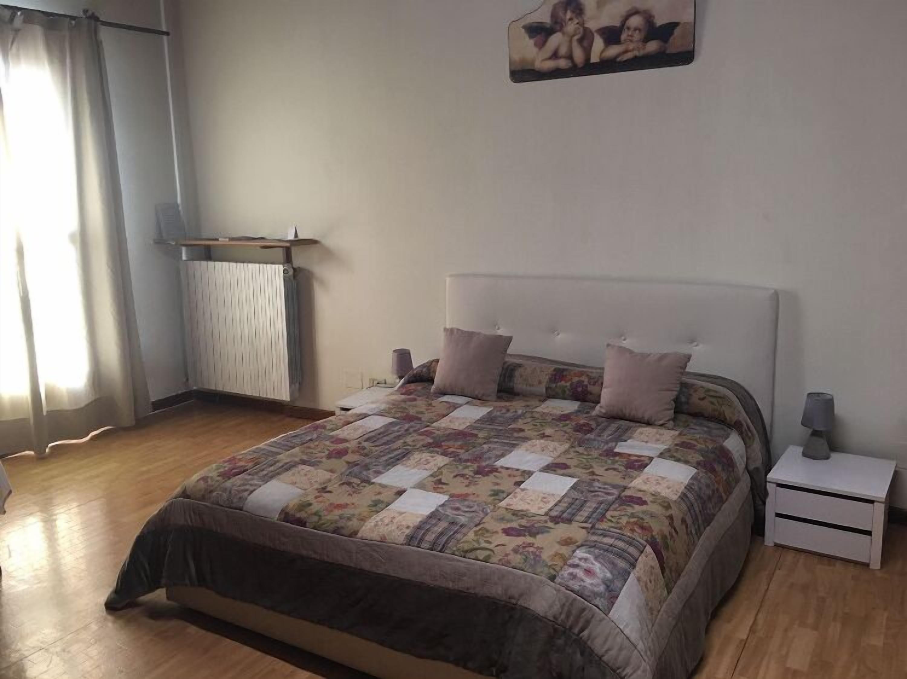 A bedroom with a large bed, a bedside table, a nightstand, a radiator, and a window with curtains.
