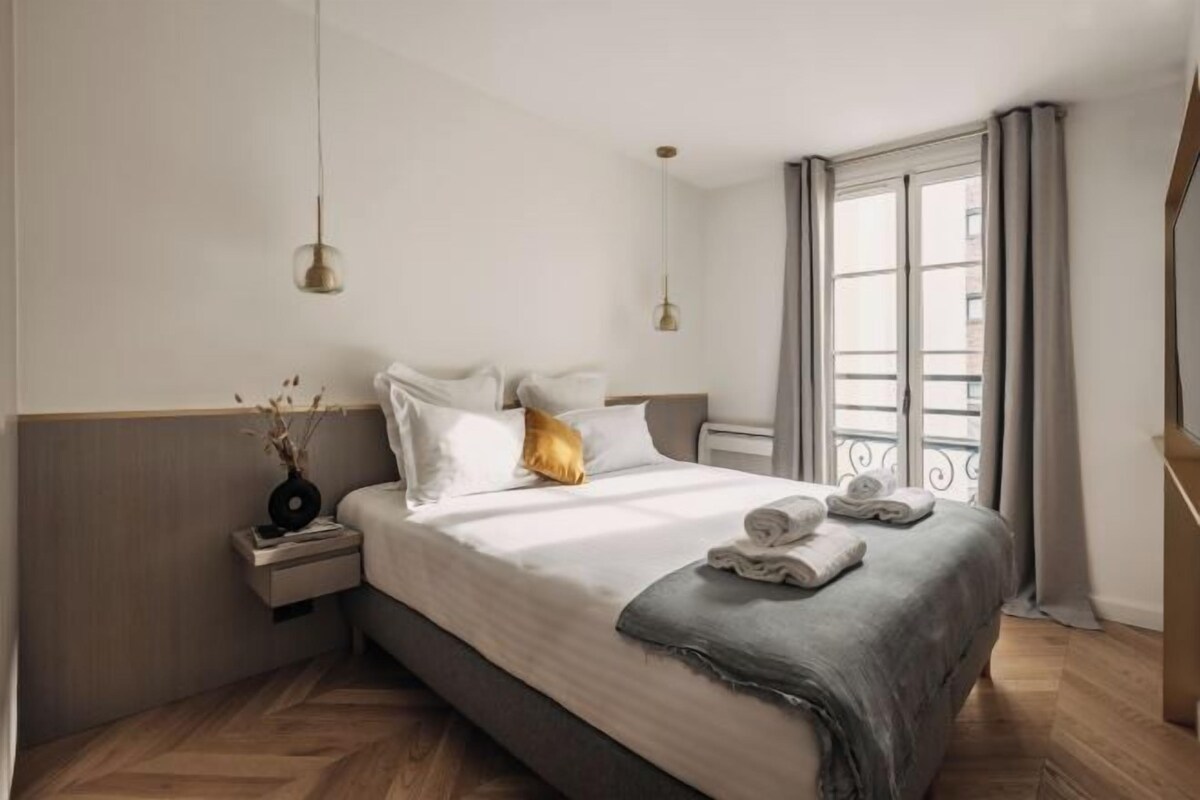 Quarto de hotel com uma cama grande, mesas de cabeceira, uma janela com cortinas e luminárias pendentes.