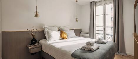 Habitación de hotel moderna con una cama grande, una mesita de noche con lámpara y vistas a la ciudad por la ventana.