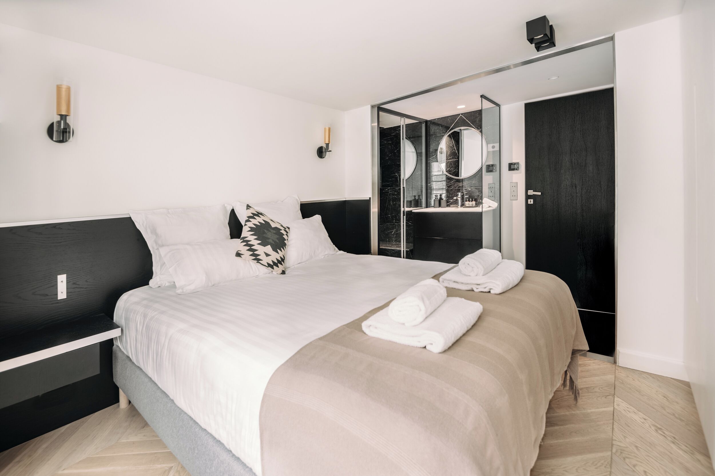 A modern hotel room with a large bed, a bedside table, a mirror, and a bathroom visible through a glass door.