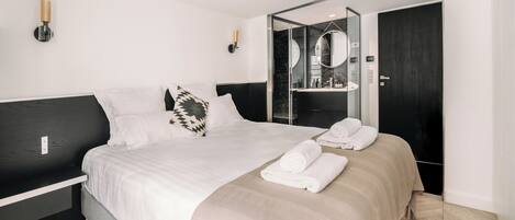 A modern hotel room with a large bed, a bedside table, a mirror, and a bathroom visible through a glass door.