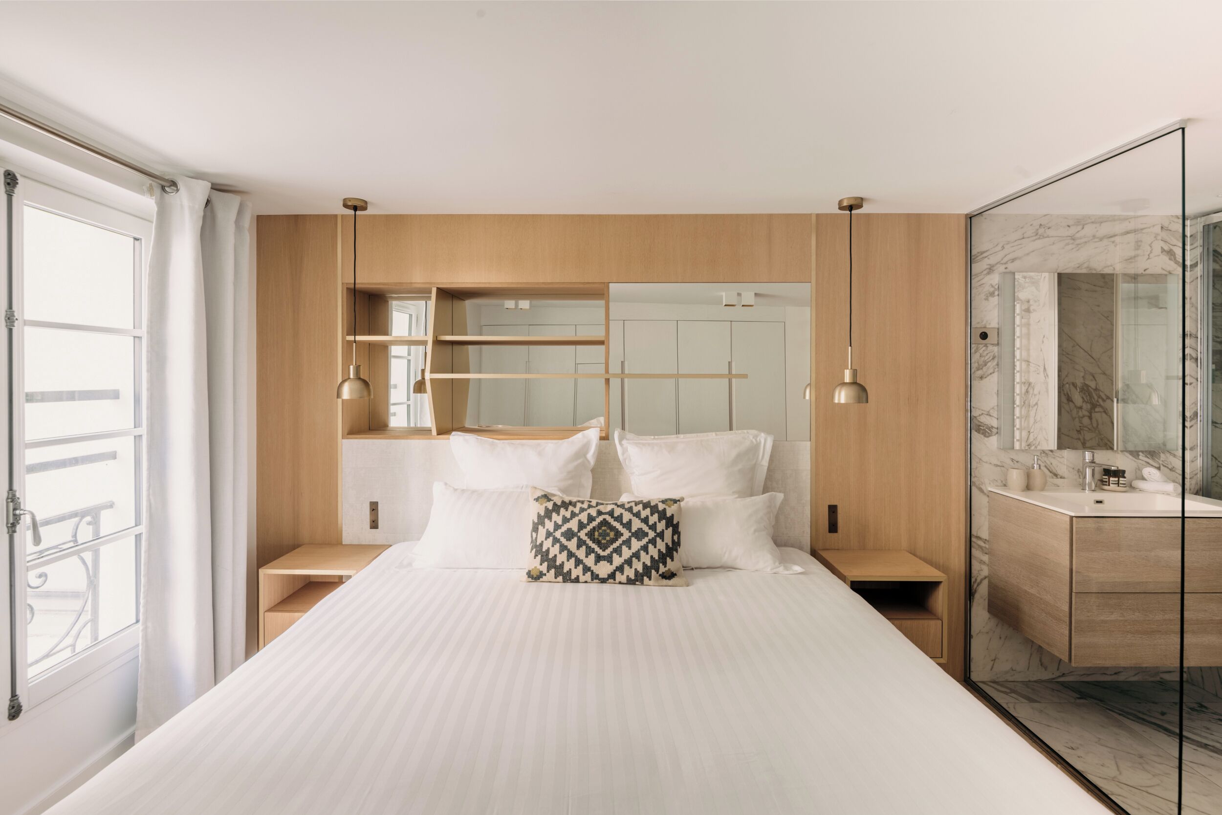 Une chambre moderne avec un grand lit, une tête de lit en bois, une table de chevet, une commode, un miroir et une salle de bain dotée d’un plan de toilette en marbre.