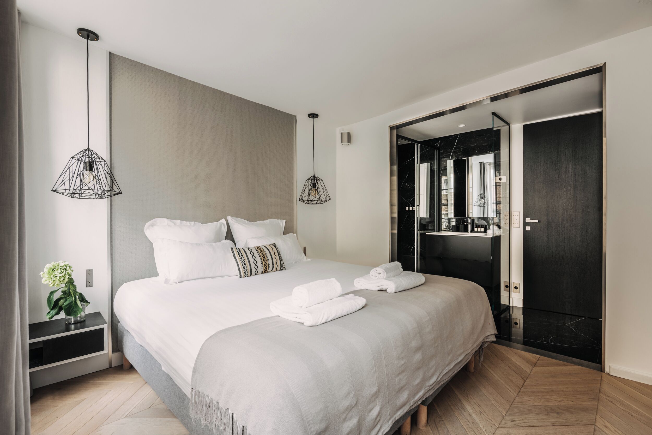 Une chambre d’hôtel moderne avec un grand lit, deux lampes suspendues, une table de chevet agrémentée d’une plante et une porte sombre donnant accès à la salle de bain.
