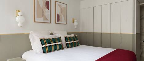 Una cama bien tendida con ropa blanca y dos almohadones decorativos, una mesita de noche con cajón y un armario con espejo.