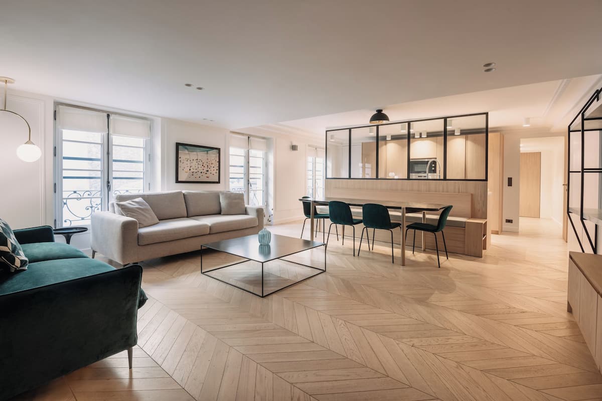 A modern living room with a sofa, a coffee table, and a dining area with chairs and a kitchen in the background.