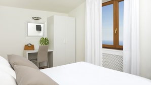 A bedroom with a bed, a desk, a chair, a plant, a window with curtains, and a view of the sea. - Villa Amiela (Massa Lubrense)