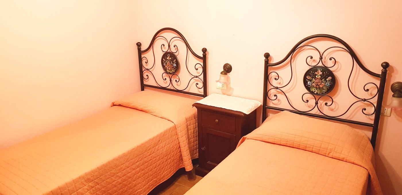 A room with two single beds with metal headboards, a small wooden nightstand, and a wall-mounted light switch.