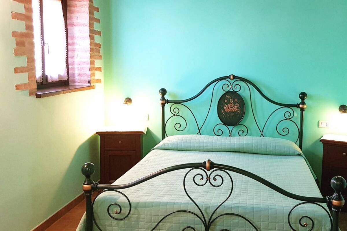 A bed with a metal frame and a quilted bedspread, two bedside tables with lamps, a window with a curtain, and a door.