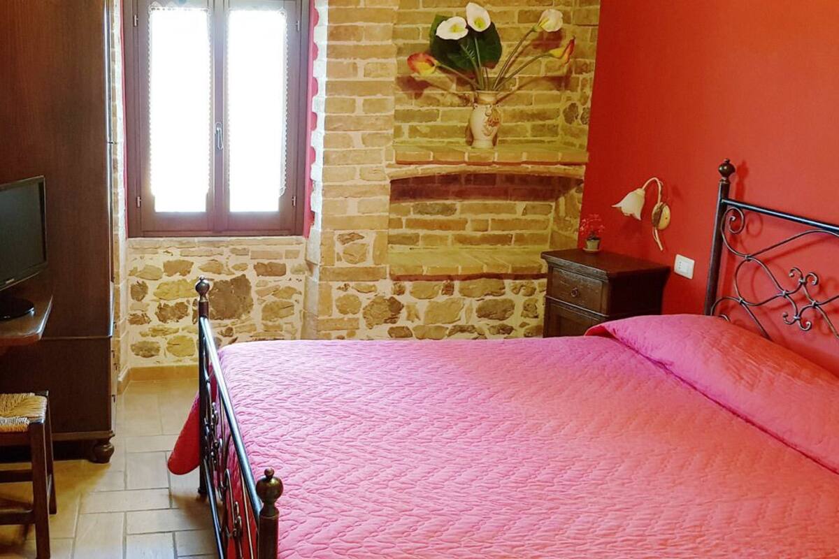 A bedroom with a brick fireplace, a bed with a pink quilt, a wooden dresser, and a television on a stand.