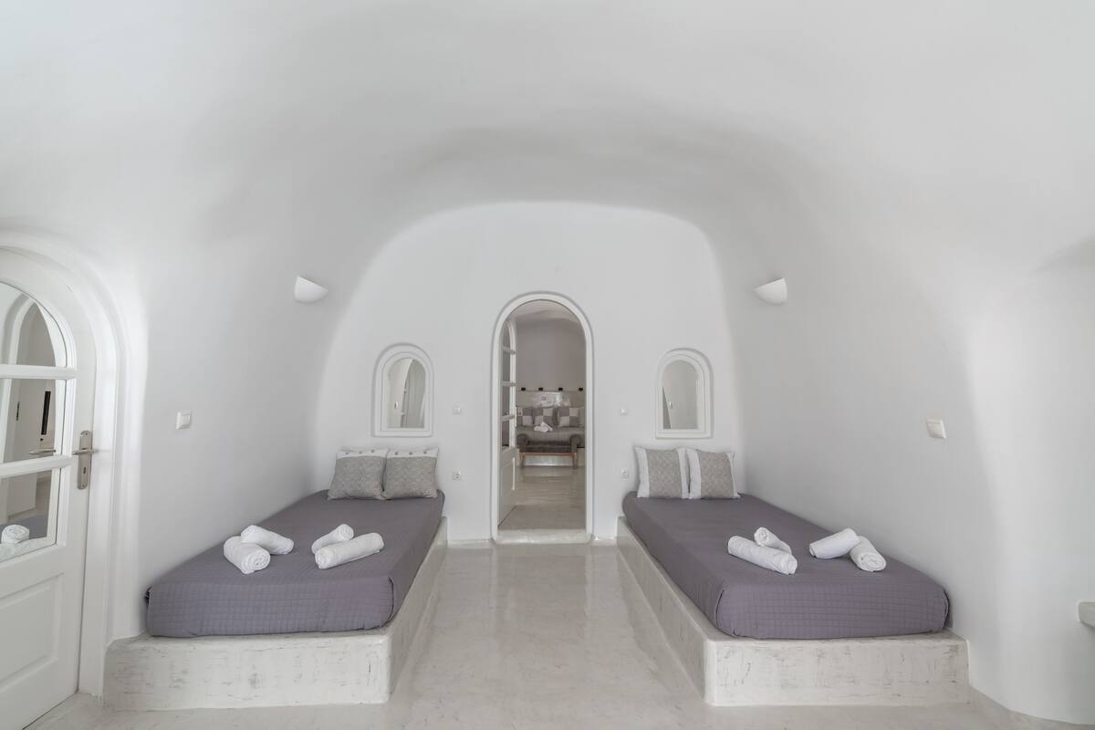 A minimalist room with two beds, a bathroom visible through an archway, and a hallway leading to another room.