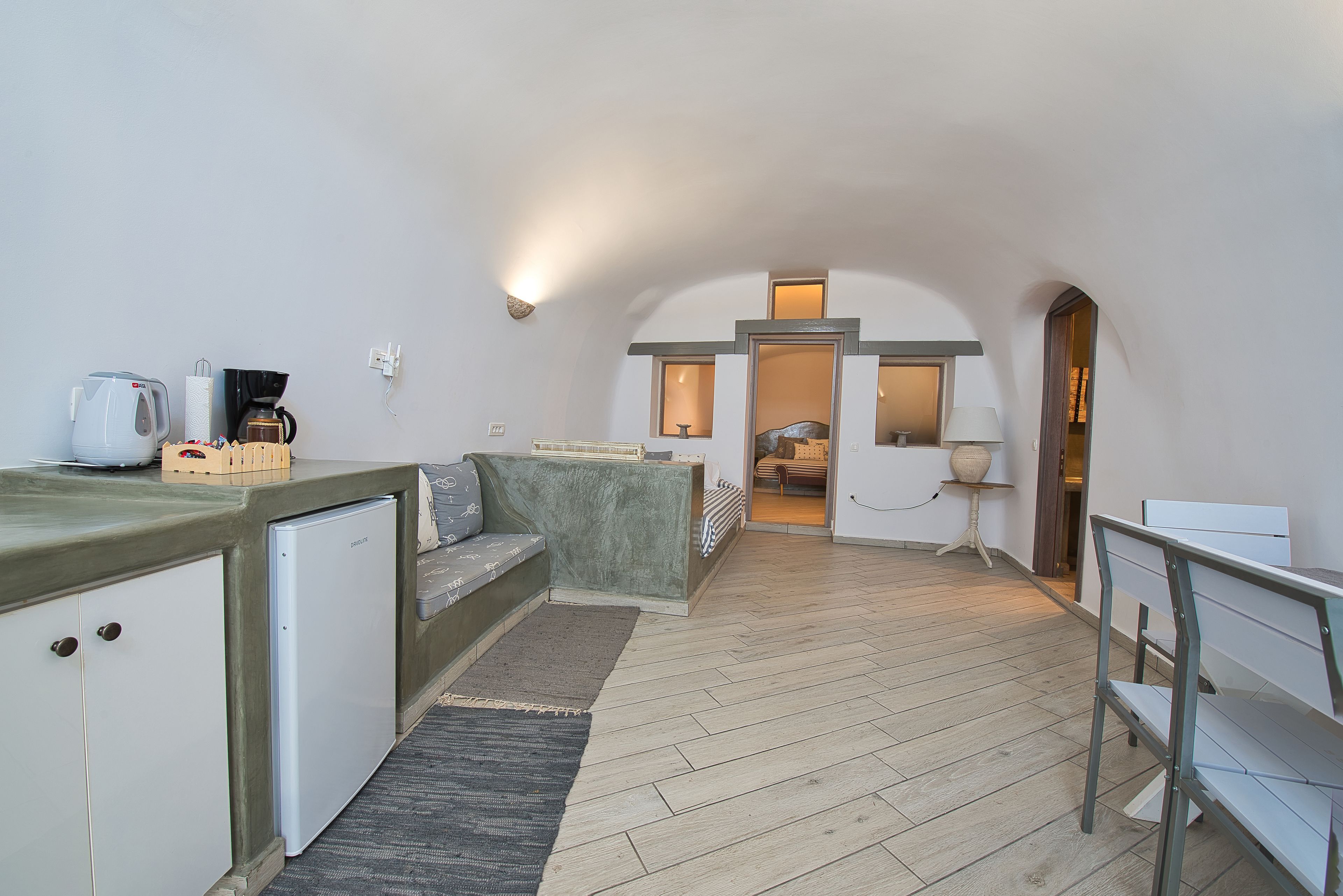 A compact kitchen with a built-in refrigerator, a coffee machine, and a small seating area with a bench and cushions.