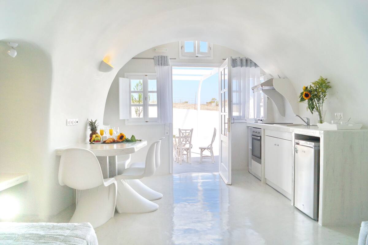 A bright, white interior with a kitchen and dining area, featuring modern furniture and a view through an open archway.