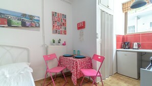 A small, compact room with a bed, a dining table with pink chairs, a refrigerator, and a kitchen area with a red backsplash. - Comfort Apartments 2 (Heraklion)