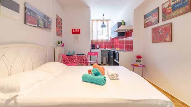 A compact hotel room with a bed, a small table with a chair, a kitchenette, and a window with blinds. - Comfort Apartments 2 (Heraklion)