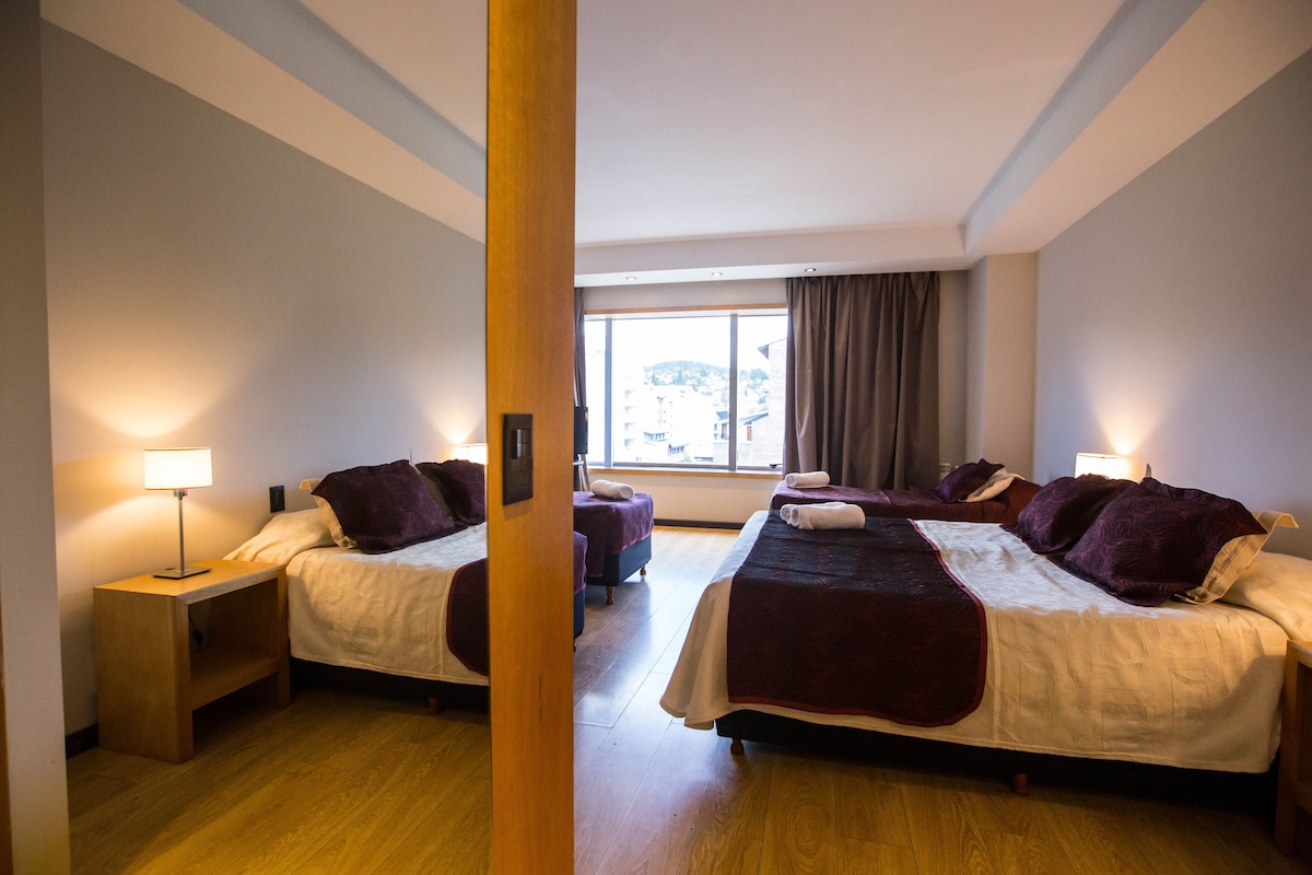 Habitación de hotel con dos camas, suelo de madera, un ventanal grande con cortinas y lámparas de noche.