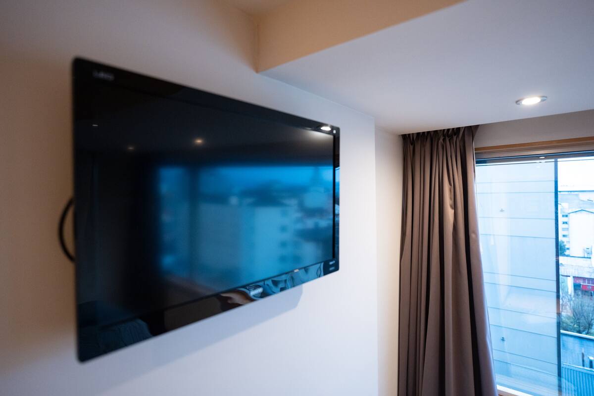 Televisor de pantalla plana montado en una pared blanca en una habitación con un ventanal y una cortina.