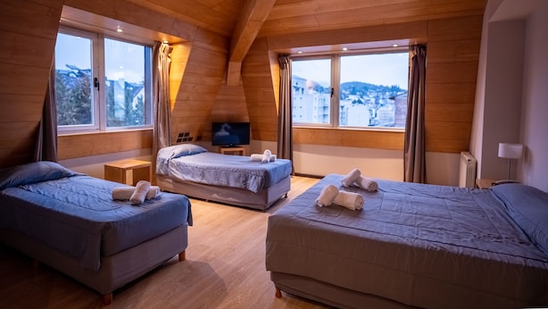 A room with three beds, wooden ceiling beams, large windows with curtains, a television on a small table, and a view of a snowy landscape. - Hotel Bariloche Suites (San Carlos de Bariloche)