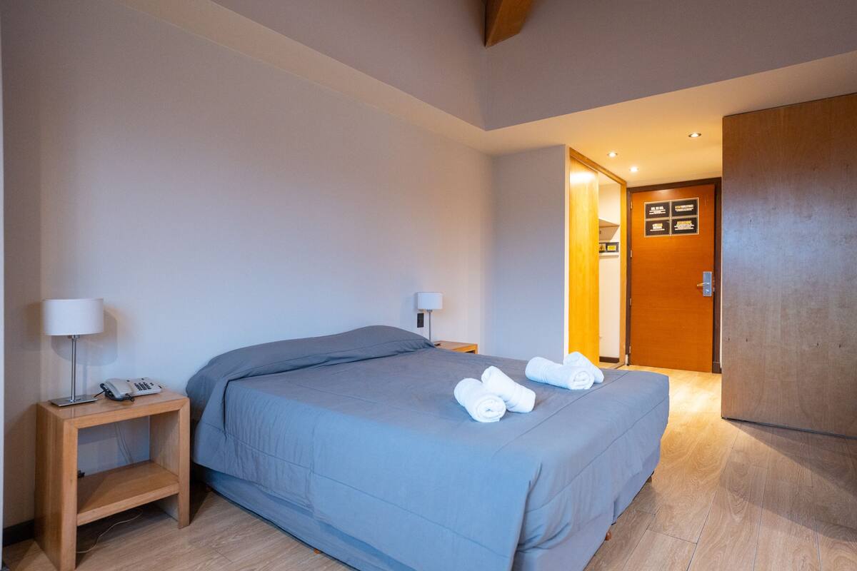 Una habitación de hotel con una cama grande, una mesita de noche con lámpara y un armario de madera.