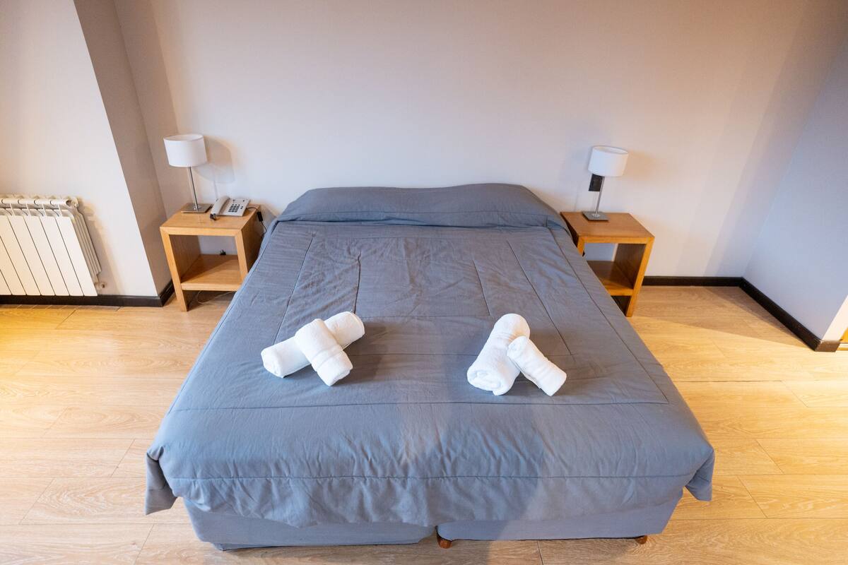 Una cama bien hecha con una colcha gris, dos toallas enrolladas sobre la cama, dos mesitas de noche con lámparas y un suelo de madera.
