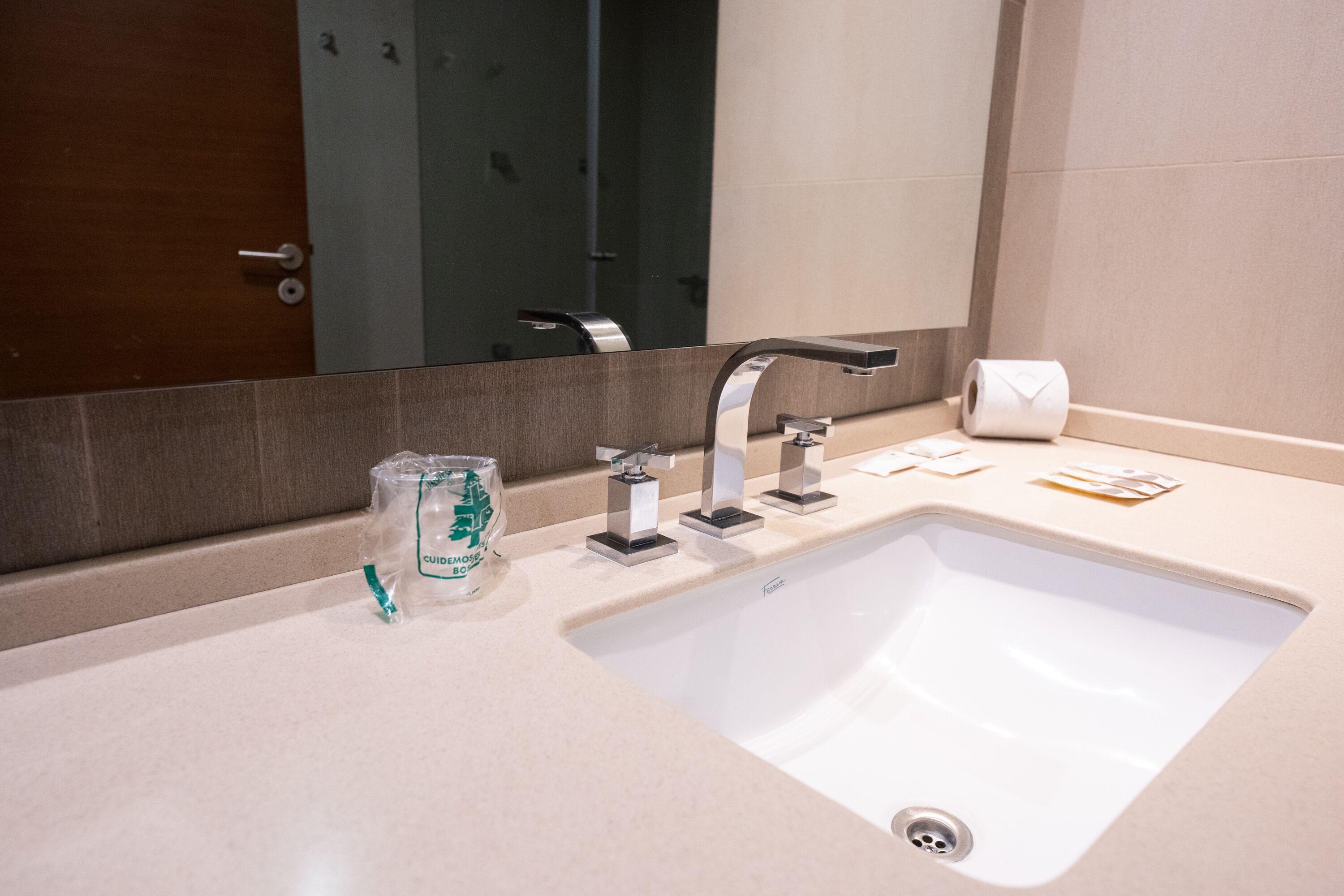 A modern bathroom with a white sink, three faucets, a mirror, and a toilet paper dispenser.