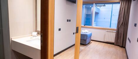 A modern hotel room with a wooden floor, a large window with curtains, a flat-screen TV mounted on the wall, and a small seating area with a blue cushion.
