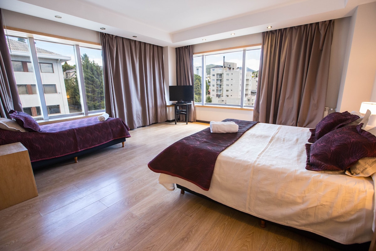 Habitación de hotel con una cama grande, sofá cama, televisor y vistas a edificios a través de amplias ventanas.