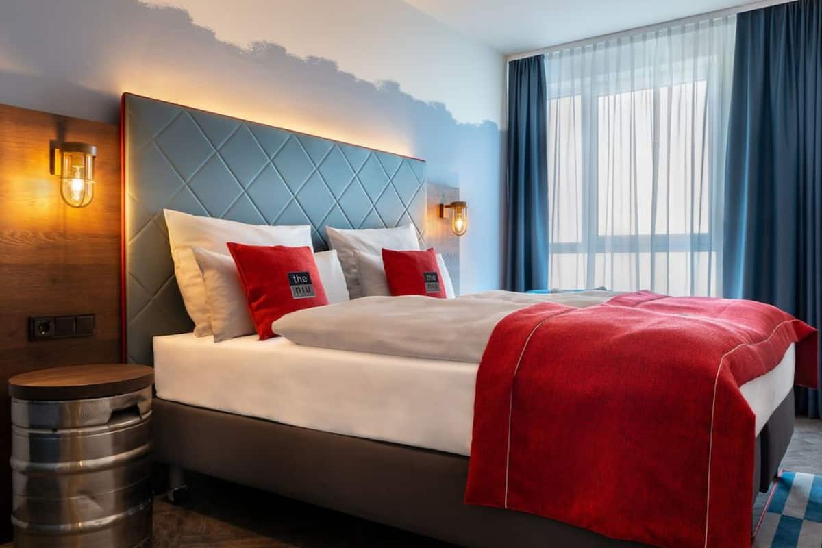 A hotel room with a large bed, a wooden headboard, two bedside lamps, and a view of the outdoors through sheer curtains.