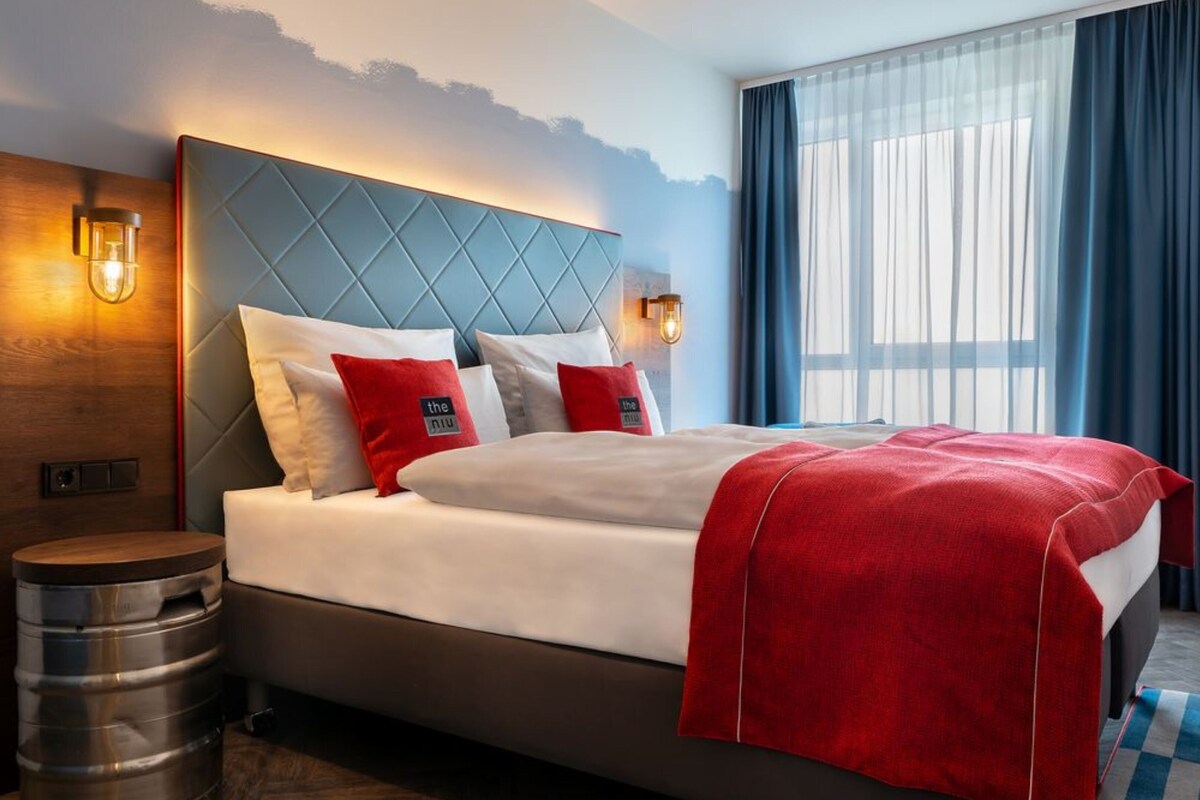 Habitación de hotel con una cama grande, cabecera de madera, dos lámparas de noche y vistas al exterior a través de cortinas translúcidas.