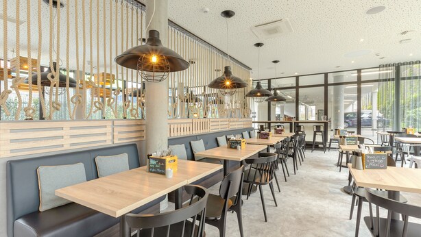 A modern restaurant interior with wooden tables, black chairs, and pendant lighting.