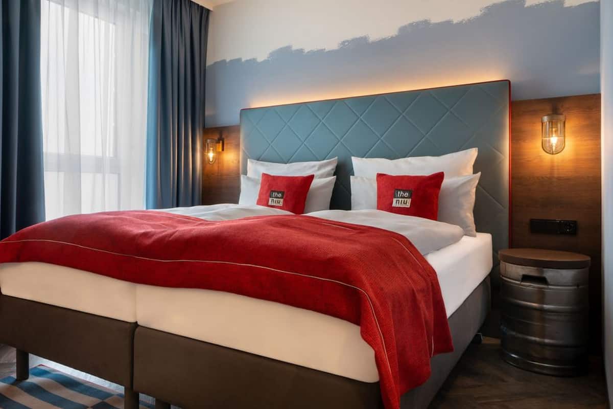 A hotel room with a large bed, two red pillows, a wooden headboard, and a nightstand with a trash can.