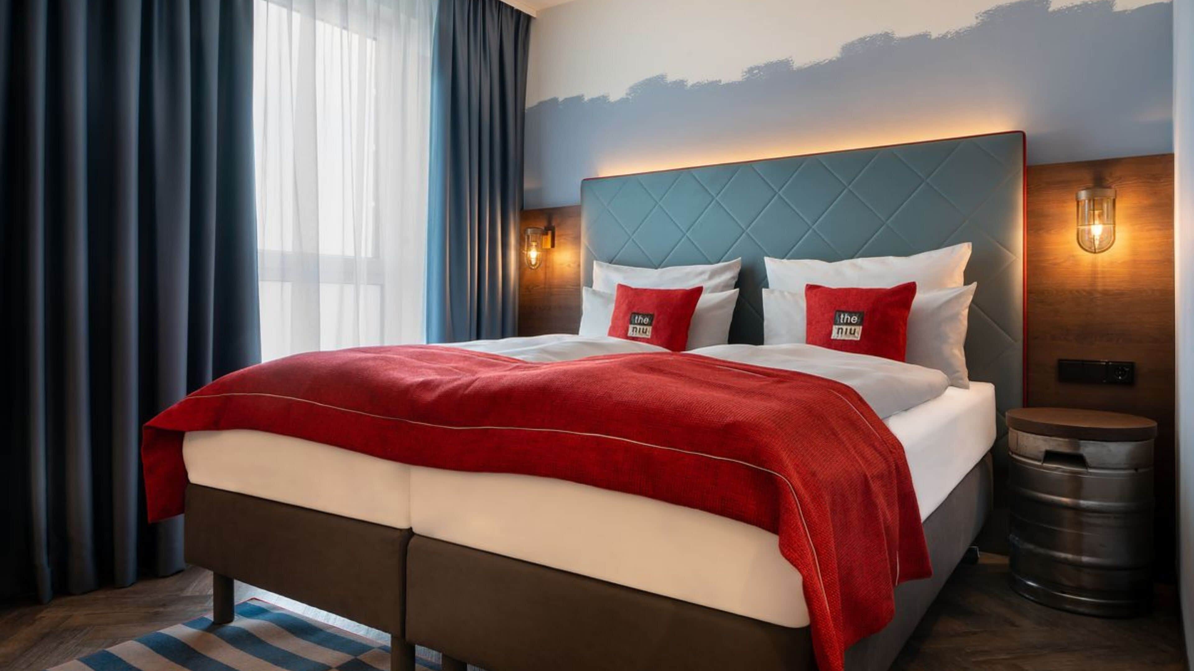 A hotel room with a large bed, two red pillows, a wooden headboard, and a nightstand with a trash can.