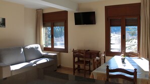 Uma sala de estar com sofá, mesa de jantar com cadeiras, televisão na parede e janelas com cortinas.