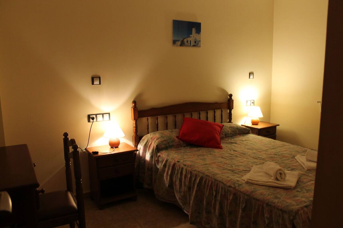 Une chambre à coucher avec une tête de lit en bois, deux tables de chevet avec des lampes, un lit recouvert d’une couvre-lit à motifs floraux et orné d’un coussin rouge, et un petit tableau accroché au mur.