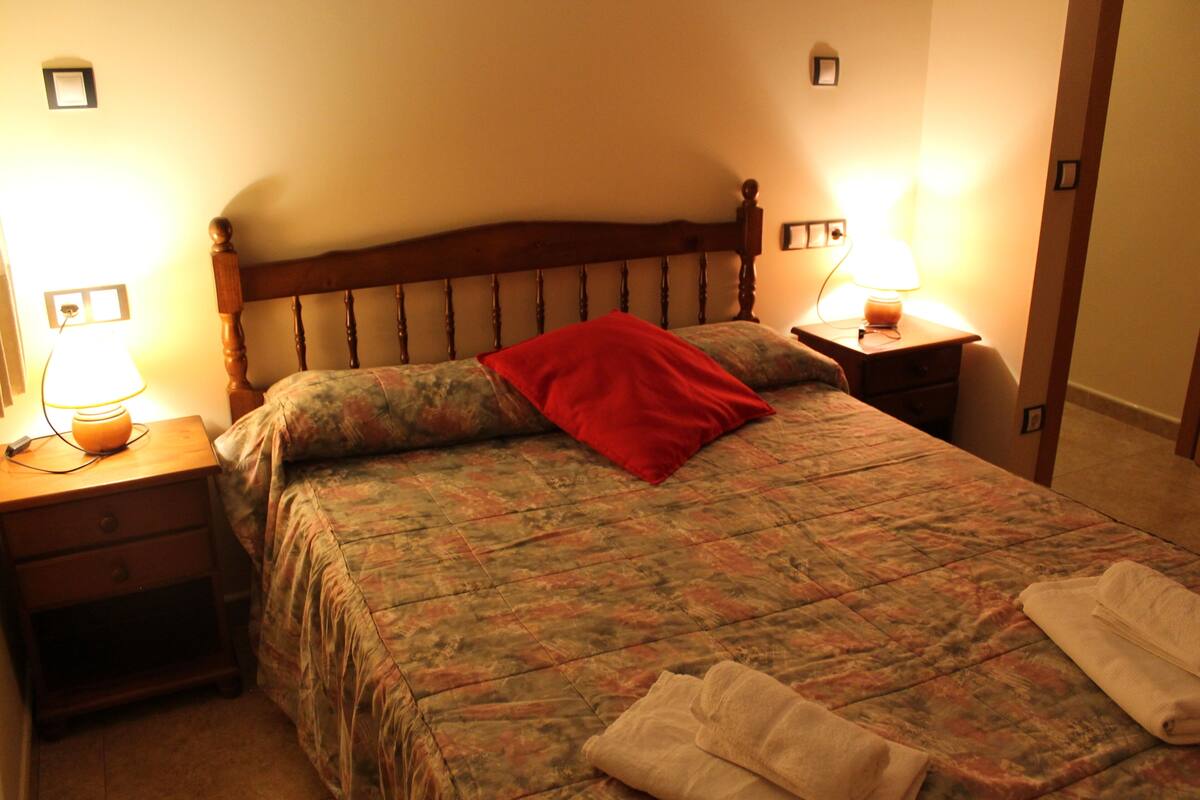 Un lit bien fait, avec une tête de lit en bois, deux tables de chevet avec des lampes et un oreiller rouge sur le lit.
