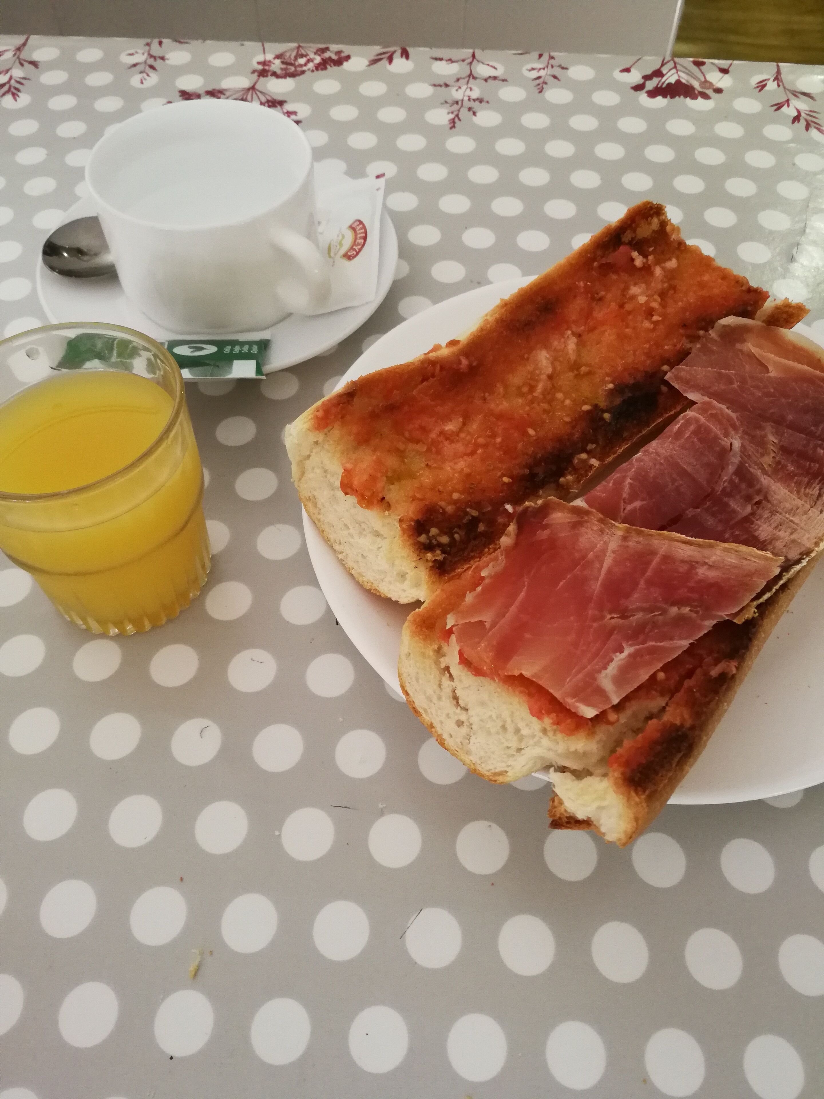A table with a polka dot tablecloth, a glass of orange juice, a white cup, and a plate with a sandwich.