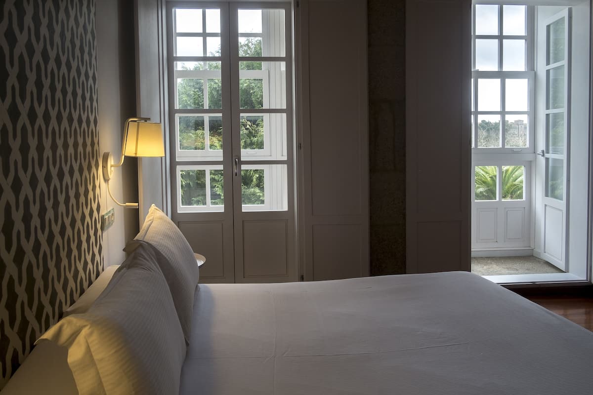 A bedroom with a bed, a nightstand, a wall-mounted lamp, and a window with a view of greenery.