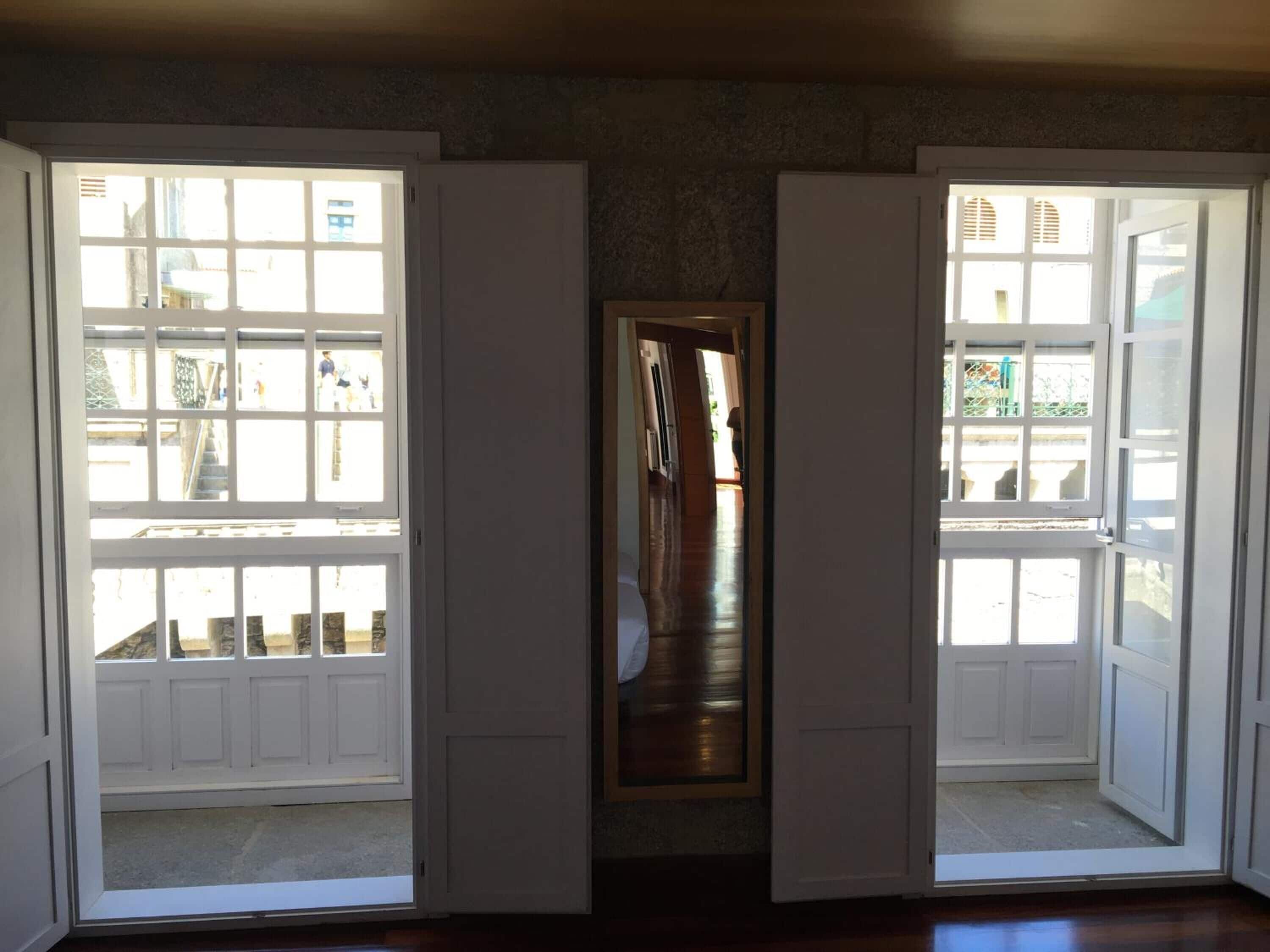 A room with two large windows, a wooden floor, and a hallway leading to another room.