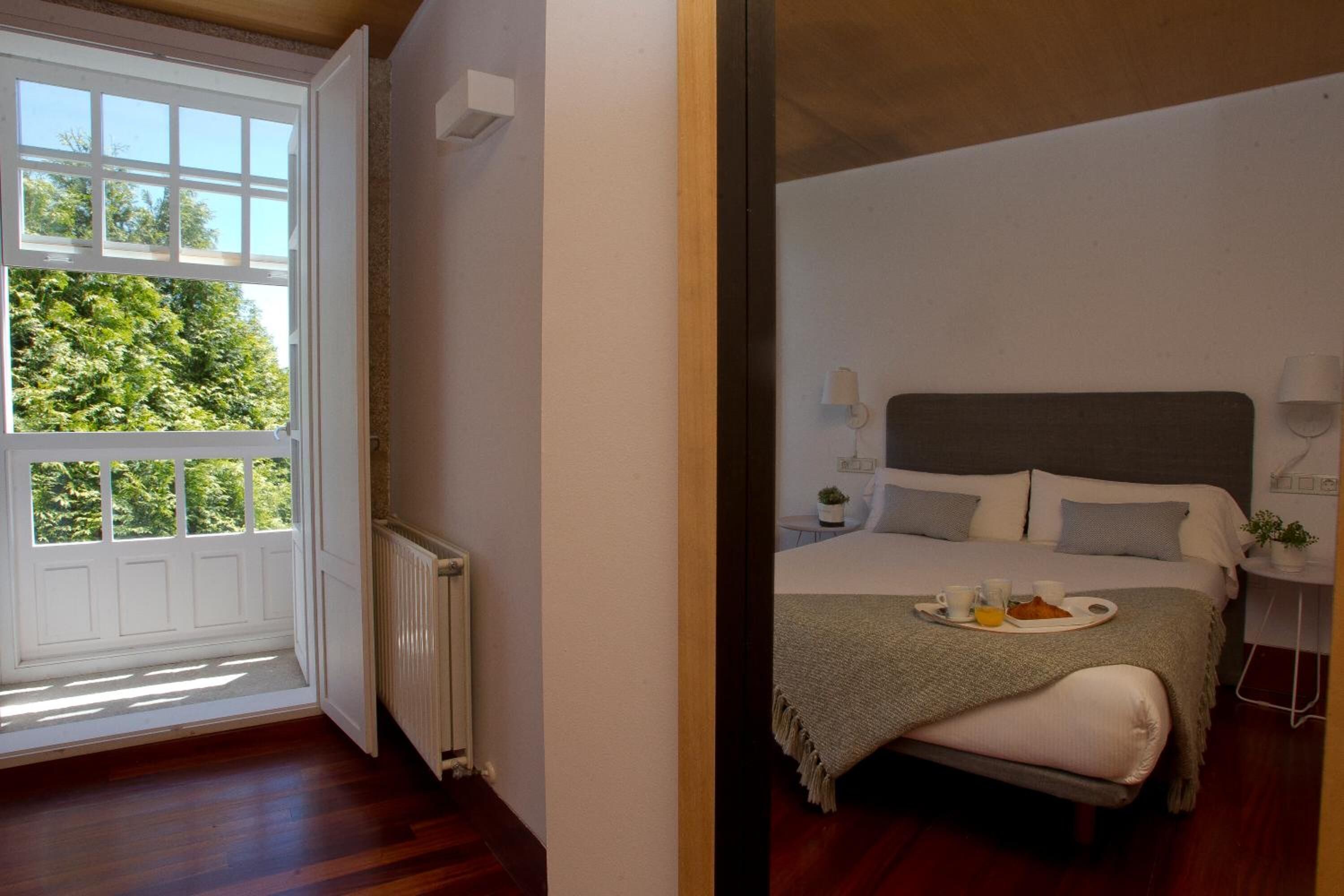 A bedroom with a large window, a bed with a tray of breakfast, and a bedside table with a lamp.