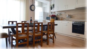 Et moderne køkken med hvide skabe, et træspisebord med stole, et ur på væggen og et vindue med gardiner.